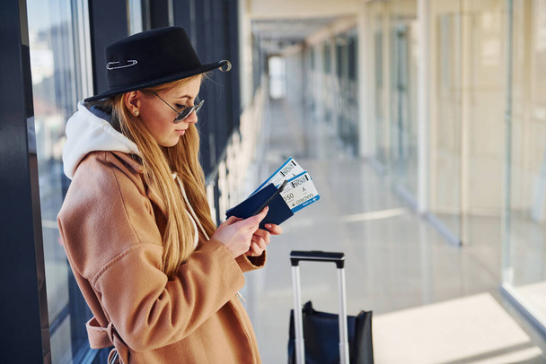 Young woman in sunglasses, warm clothes and hat standing indoors in airport with phone and tickets in hands. - Photo, Image