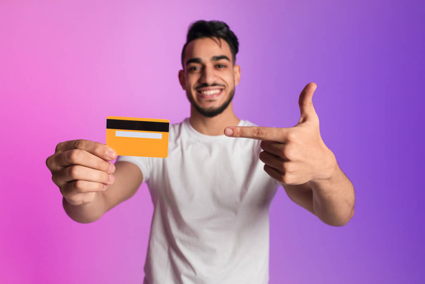 Happy young Arab man pointing at credit card, promoting electronic money in neon light, selective focus - Φωτογραφία, εικόνα