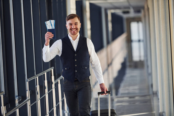 Young male passenger in elegant formal clothes is in the airport hall showing his tickets and smiling. - Fotoğraf, Görsel