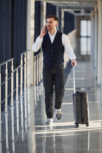 Young male passenger in elegant formal clothes is in the airport hall walking with baggage and talking by phone. - Фото, изображение