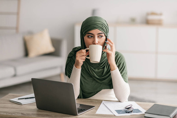 Νεαρή Άραβα γυναίκα με μαντίλα που εργάζεται στο διαδίκτυο στο γραφείο στο σπίτι, χρησιμοποιεί φορητό υπολογιστή, τηλεφωνεί στο smartphone και πίνει καφέ - Φωτογραφία, εικόνα