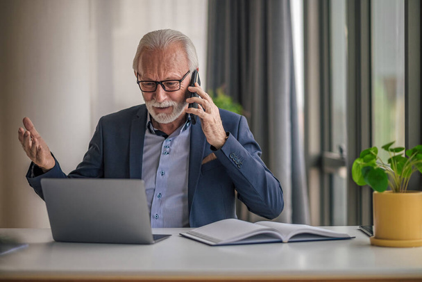 Aggódó üzletember, aki mobilon beszél, miközben a laptopon dolgozik. Az aggódó, idősebb férfi szakember formális ruhát visel. Az irodájában ül az asztalnál.. - Fotó, kép