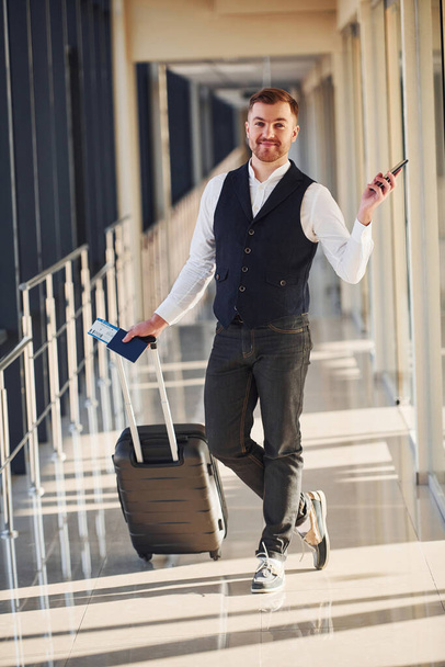 Male passenger in elegant formal clothes is in the airport hall with baggage, tickets and phone. - Fotoğraf, Görsel