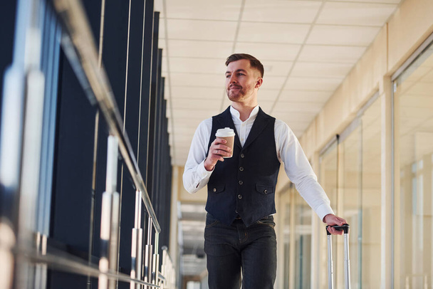 Young male passenger in elegant formal clothes is in the airport hall walking with baggage and cup of drink. - Fotografie, Obrázek