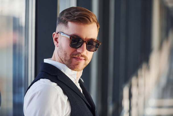 Man in elegant clothes and sunglasses is standing indoors and posing for the camera. - Foto, imagen