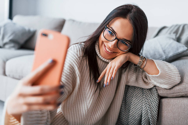 Having conversation. Young beautiful woman in glasses sitting at home alone with phone in hands. - Foto, imagen