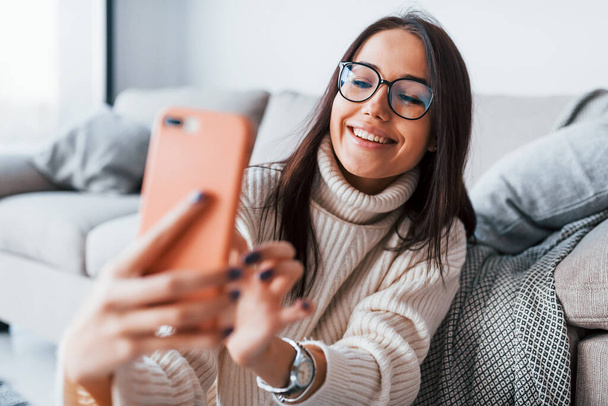 Making selfie. Young beautiful woman in glasses sitting at home alone with phone in hands. - Foto, Imagem