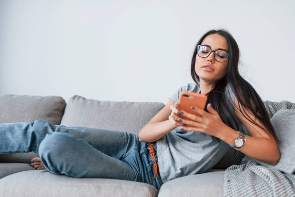 Young beautiful woman in casual clothes lying down at home alone with smartphone in hands. - Foto, afbeelding
