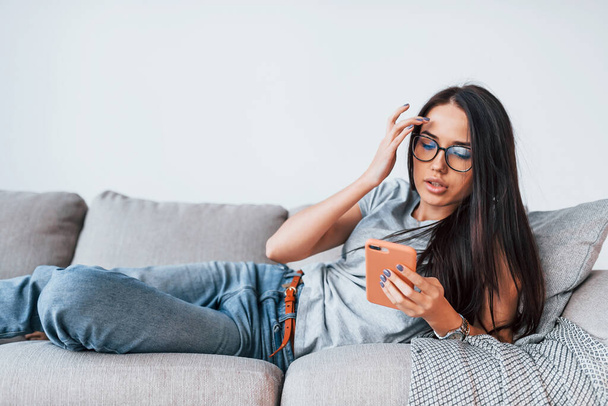 Young beautiful woman in casual clothes lying down at home alone with smartphone in hands. - Foto, imagen