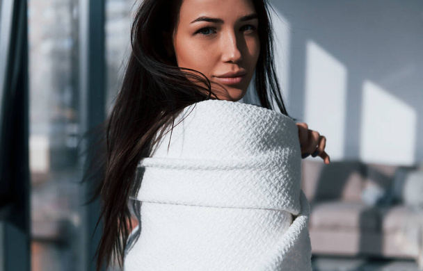 Young beautiful brunette standing indoors and covering her body by white towel. - Фото, зображення