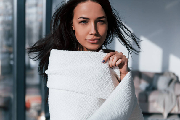 Young beautiful brunette standing indoors and covering her body by white towel. - Foto, Imagen