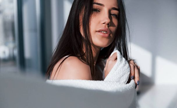 Young beautiful brunette standing indoors and covering her body by white towel. - Zdjęcie, obraz