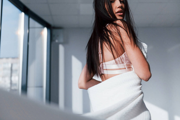 Young beautiful brunette standing indoors and covering her body by white towel. - Φωτογραφία, εικόνα