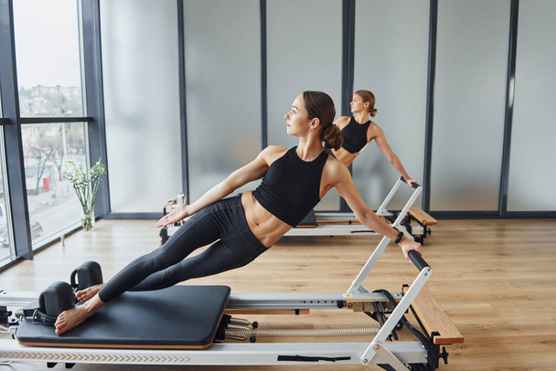Standing on gym equipment and doing stretches. Two women in sportive wear and with slim bodies have fitness yoga day indoors together. - Photo, Image