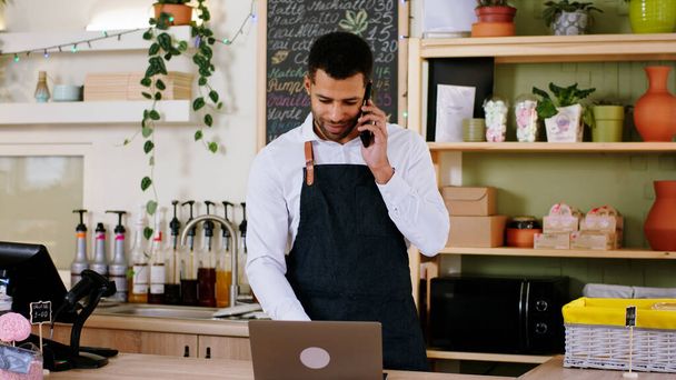 Concept of entrepreneur and small business Afro American barista guy very charismatic in the cozy coffee shop typing on the laptop the new order while speaking with the customer on the smartphone - 写真・画像