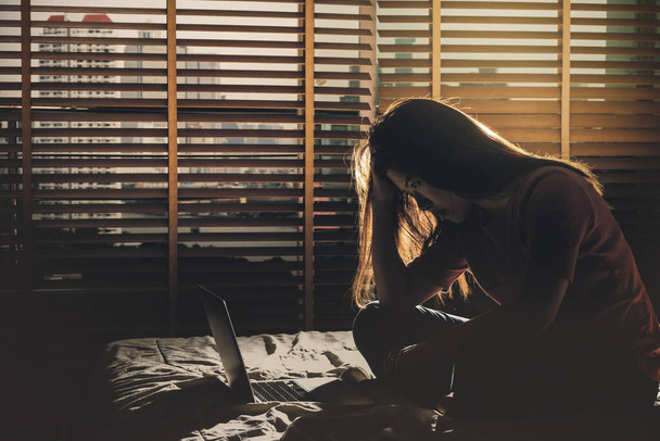donna depressa seduta testa in mano quando si lavora con la tecnologia portatile sul letto nella camera da letto buia con ambiente a bassa luce, concetto drammatico
 - Foto, immagini