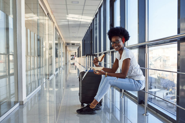 Young african american female passanger in casual clothes is in airport with baggage eating some food. - Fotografie, Obrázek