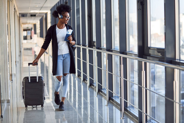 Young african american female passanger in casual clothes and headphones is in airport with baggage. - Φωτογραφία, εικόνα