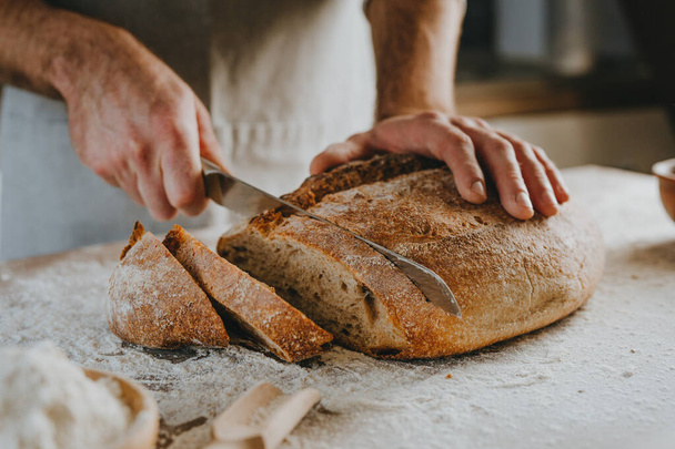 Unrecognizable young man in apron cutting homemade whole grain bread. Selective focus. - Foto, Imagem
