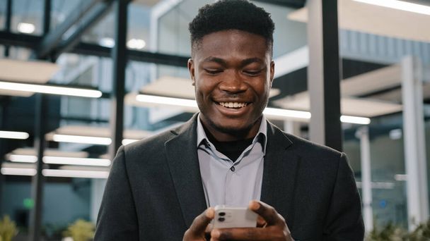 Onnellinen hymyilevä afrikkalainen kaveri amerikkalainen mies liikemies pomo työntekijä tilalla moderni langaton laite tekstiviesti älypuhelimeen sosiaalisen median sovellukset selailevat internetin pelaaminen mobiilipeli toimistossa - Valokuva, kuva