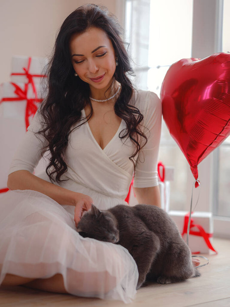 Hermosa joven feliz en vestido blanco y gato gris. fiesta de vacaciones. Modelo alegre posando con globos en forma de corazón rojo, divirtiéndose, celebrando el Día de San Valentín. Símbolo del amor - Foto, imagen