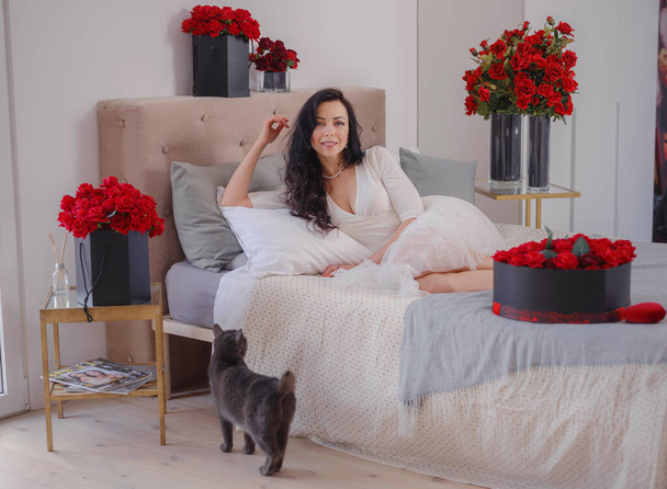 mladá žena v bílých dresech sedí na posteli s šedou kočkou. Ranní milostná ložnice. Výročí, Všechno nejlepší, Svatba, Den lásky, Den žen, Valentýna a pozitivní emoce - Fotografie, Obrázek