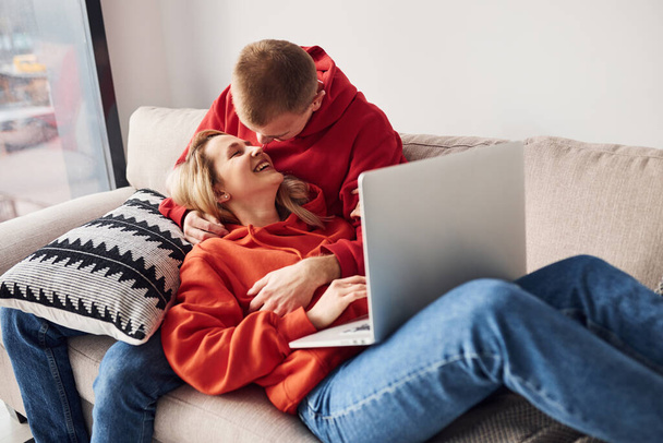 Using laptop that is on the legs. Young lovely couple together at home spending weekend and holidays together. - Photo, image