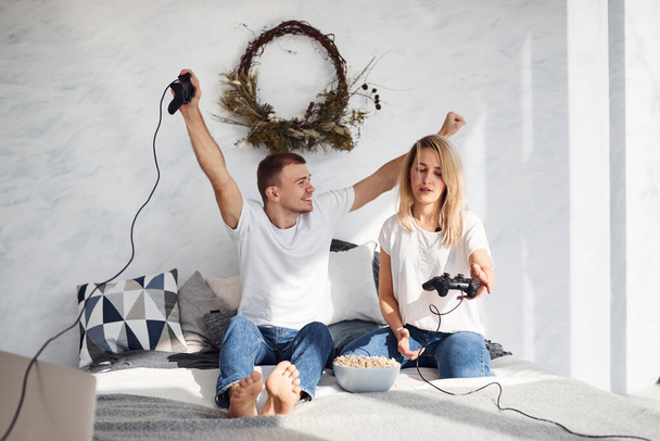 Young lovely couple together at home playing video games on bed with popcorn at weekend time. - Foto, afbeelding