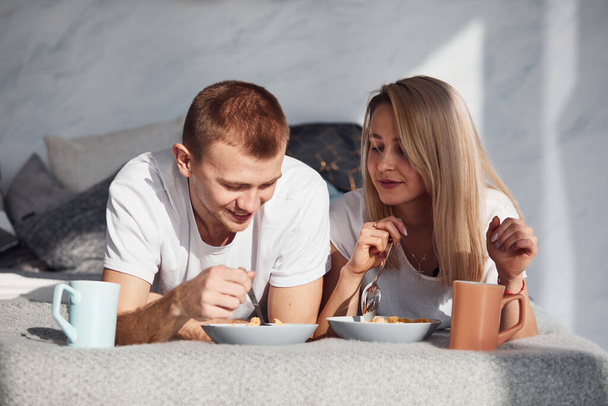 Young lovely couple have a breakfast at home while lying down on bed. - Фото, изображение