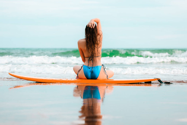 όμορφο κορίτσι από την Ασία που φοράει μπικίνι κάθεται στη σανίδα του σερφ και ποζάρει στην παραλία της άμμου, σε μια τροπική τοποθεσία, ταξιδεύουν καλοκαιρινές διακοπές χαλαρώστε στην έννοια της θάλασσας - Φωτογραφία, εικόνα