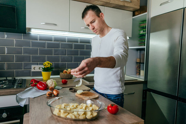 Mies leikkaa sipulia ja laittaa sen paistoastiaan. Tunnistamaton mies keittää vihanneksia keittiössä. Lähikuva miehillä kädet valmistelee perunaa ja sipulia kokki. Valikoiva painopiste. - Valokuva, kuva