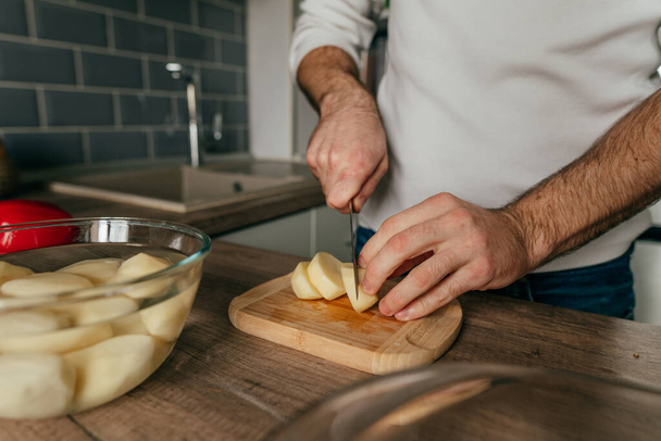 Człowiek krojący ziemniaki na tablicy. Nierozpoznawalny człowiek gotujący warzywa w kuchni. Zbliżenie męskich rąk przygotowujących ziemniaki do gotowania. Skupienie selektywne. - Zdjęcie, obraz