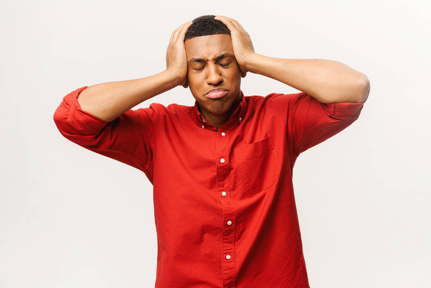 Upset young African-American short-cutted guy in red shirt holds head with both hands and suffers from strong headache - Фото, изображение