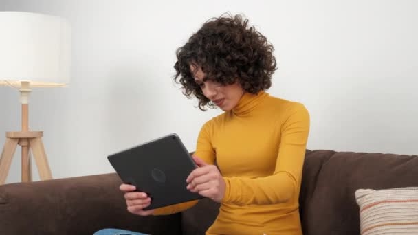 Kręcone kobieta emocjonalnie gra w gry wideo na tablecie, siedzi na kanapie w domu - Materiał filmowy, wideo