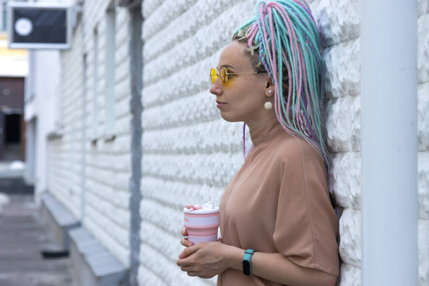 Young modern woman with colored dreadlocks hairstyle with a glass of coffee for a walk, stands near a street wall. - Фото, изображение