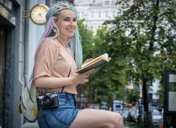 Young woman traveler with a backpack and retro photo camera. Travel and read book out sightseeing in a history city. Student woman, vacation. Learning foreign languages in a different country.  - Zdjęcie, obraz