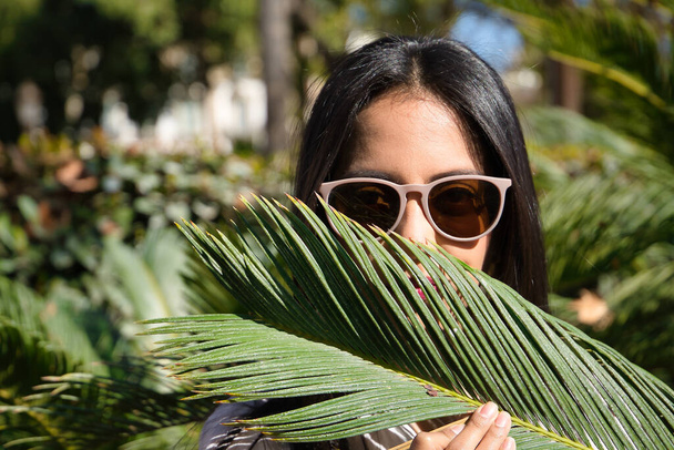 Portrait of beautiful young woman, South American, with sunglasses, covering her face with a green palm leaf. Concept beauty, fashion, plants, nature, travel, green. - 写真・画像
