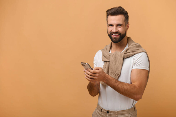 Portrait of cheerful brunette man with beard in white t-shirt using cellphone and smiling, reading good news message, enjoying mobile application. Man looking at the camera with smile - Foto, Bild