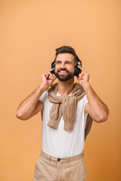 Relax, favorite music, fun and pleasure at spare time. Smiling excited student man in casual with modern headphones enjoying of the music isolated on orange - Valokuva, kuva