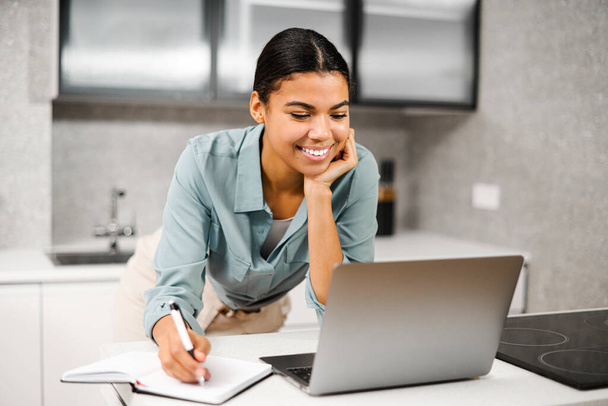 Entrepreneur young woman using laptop in home office. African-american woman concentrated looks at computer screen and making notes - 写真・画像