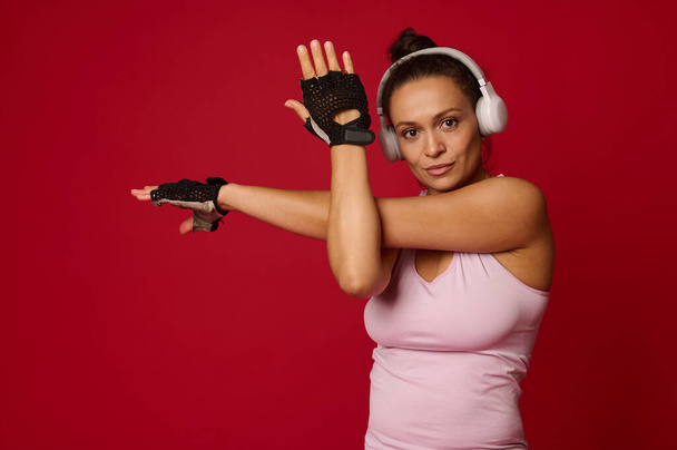 Confident active middle-aged active multi-ethnic woman in sportswear and headphones stretching arms, looking at camera against red background with copy ad space - Photo, image