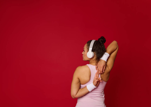 Vista trasera de una atleta femenina multiétnica determinada con auriculares que se extienden los brazos mientras se ejercita contra el fondo de color rojo con espacio de copia de publicidad - Foto, imagen