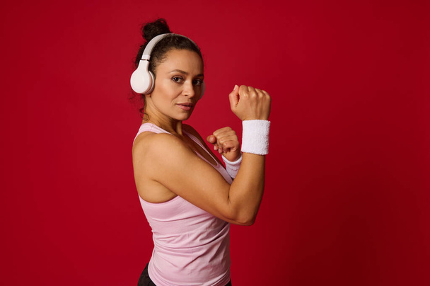 Ελκυστική αυτοπεποίθηση αποφασισμένη εκπληκτική Ισπανίδα αθλήτρια φορώντας ακουστικά σφίγγοντας τις γροθιές της ενώ στέκεται σε στάση μάχης σε κόκκινο φόντο με χώρο αντιγραφής - Φωτογραφία, εικόνα