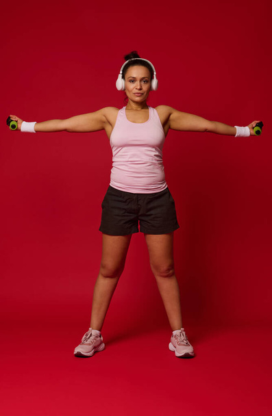 Volledige lichaamsomvang weergave van vrouwelijke atleet oefenen met halters tegen rode achtergrond met kopieerruimte voor reclame tekst. Sport, fitness, gezonde en actieve levensstijl, gewichtsverlies concept - Foto, afbeelding