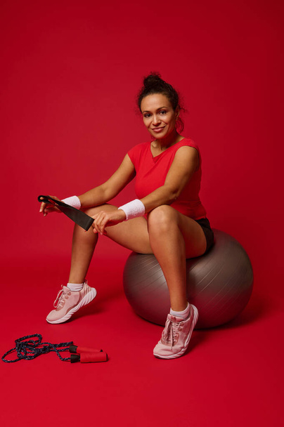 Hispanic kaunis hymyilevä urheilullinen nainen tilalla elastinen kunto bändi ja istuu kunto pallo harjoittelun jälkeen eristetty punaisella taustalla. Käsitys laihuudesta, terveellisistä elämäntavoista ja harjoituksesta. - Valokuva, kuva