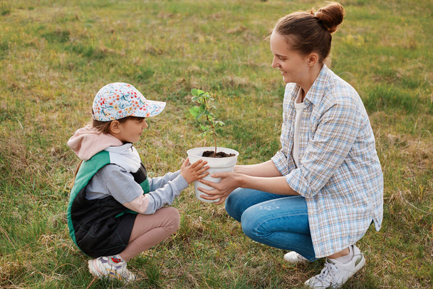 Full length profile portrait of kind little girl holding pot with raspberry seedlings together with mommy, smiling cute gardeners being going to plant tasty berry bush. - Foto, Imagem