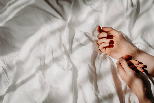 Elegant hands lie on the white bed sheet in the sunlight. Bed with white linens. The concept of a good morning, stress relief, self-care, relaxation and time for yourself - Zdjęcie, obraz