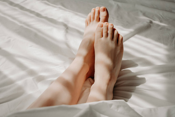 Woman is lying with bare feet in bed, sleeping under white cosy and comfortable bedclothes. Female feet on bed. Good morning, woman sexuality concept, self-care and relaxation, resting concept - Foto, afbeelding