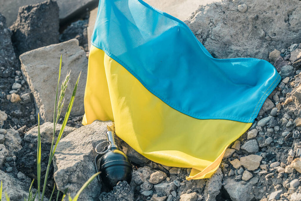 Molozların üzerinde yatan Ukrayna bayrağı, yerde yatan bir el bombasının yanında, silahlı saldırı ve savaş kavramı. - Fotoğraf, Görsel