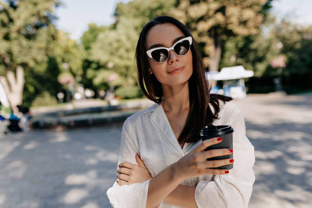 Elegant stylish woman with straight dark hair wearing sunglasses and white shirt is holding cup with coffee and posing at camera in sunlight in green summer park in the city. High quality photo - Photo, Image